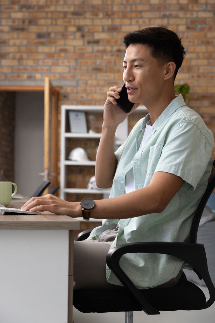 Asian Man Typing at Desk While Talking on Smartphone in Office