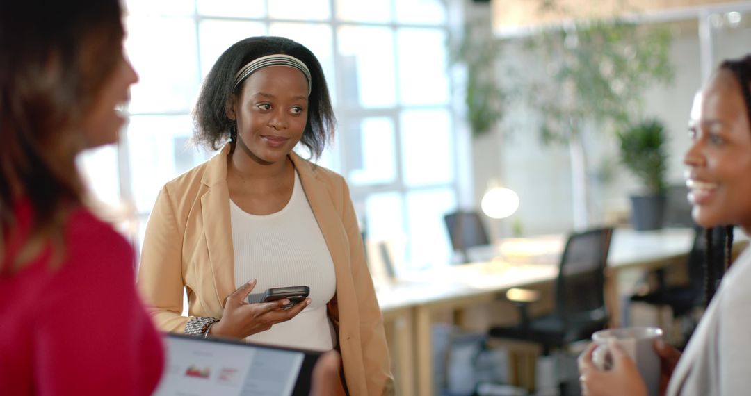 Diverse female coworkers collaborating with phone, tablet and coffee in bright open-plan office