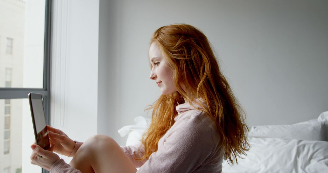 Young woman sitting by window using tablet in morning light - Free Images, Stock Photos and Pictures on Pikwizard.com