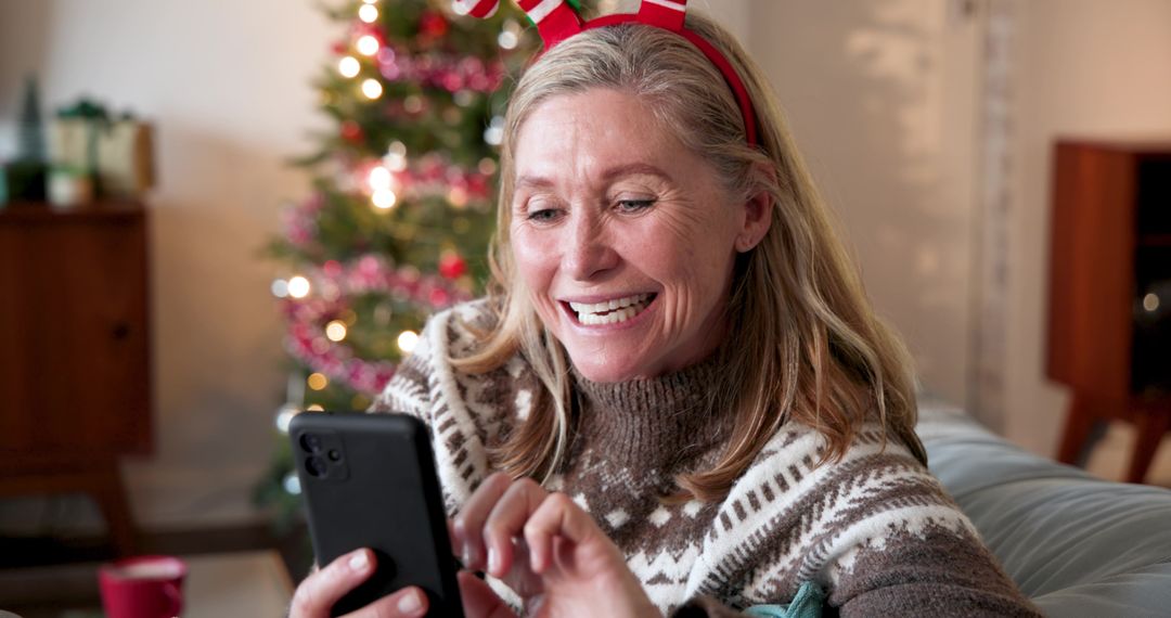 Smiling Woman with Antler Headband Using Smartphone Near Christmas Tree