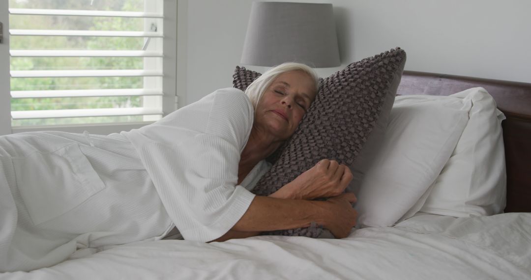 Senior woman cuddling pillow while napping on bed in cozy modern bedroom - Free Images, Stock Photos and Pictures on Pikwizard.com