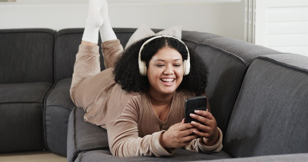 Smiling woman lounging on gray sofa, listening with headphones and using smartphone