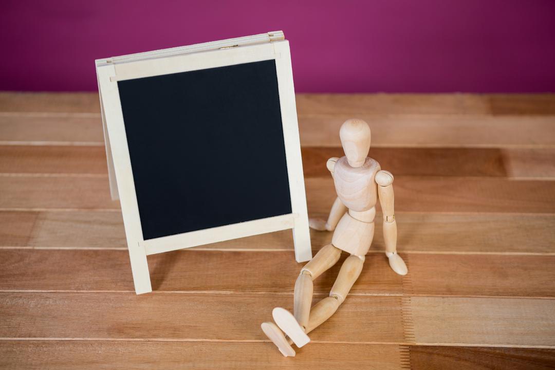 Wooden Figurine Sitting Near Blank Chalkboard on Wooden Floor - Free Images, Stock Photos and Pictures on Pikwizard.com