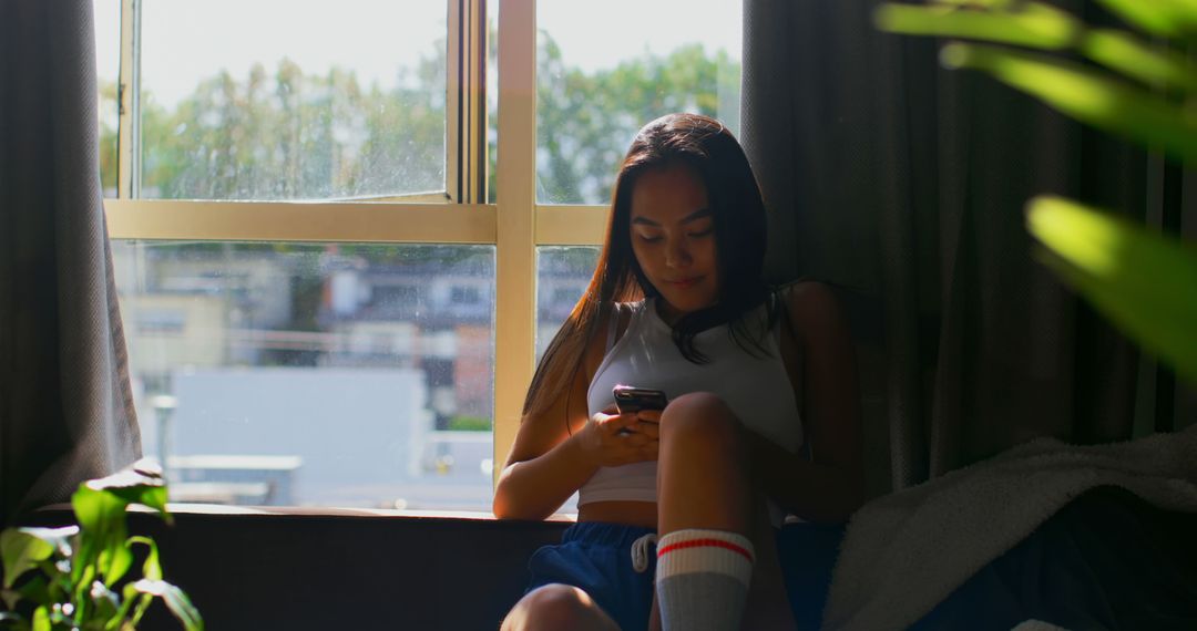 Young Woman Relaxing by Window with Smartphone on Sunny Day - Free Images, Stock Photos and Pictures on Pikwizard.com