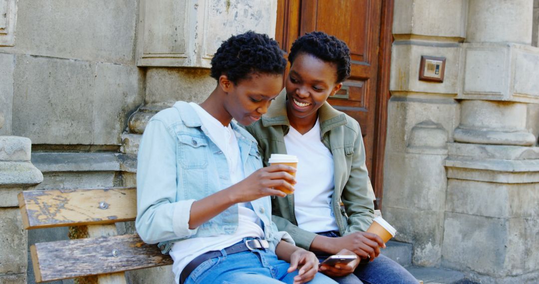 Smiling African American Twin Sisters Sharing Coffee and Smartphone in Urban Setting