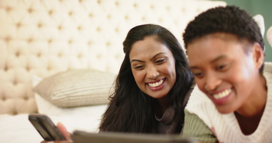Diverse Female Friends Smiling While Using Gadgets in Cozy Bedroom