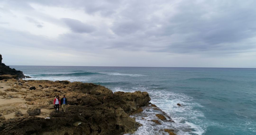 Couple Standing on Rocky Shoreline Looking at Ocean Waves Under Cloudy Sky - Free Images, Stock Photos and Pictures on Pikwizard.com