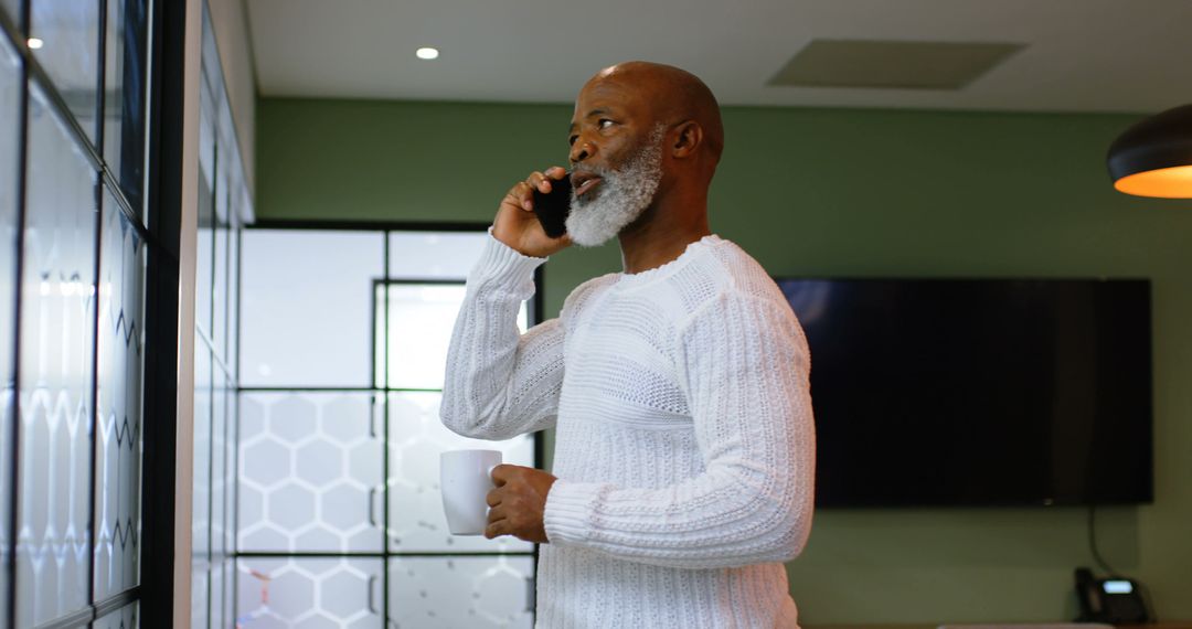 Mature Man Speaking on Cellphone Holding Coffee in Modern Living Room - Free Images, Stock Photos and Pictures on Pikwizard.com