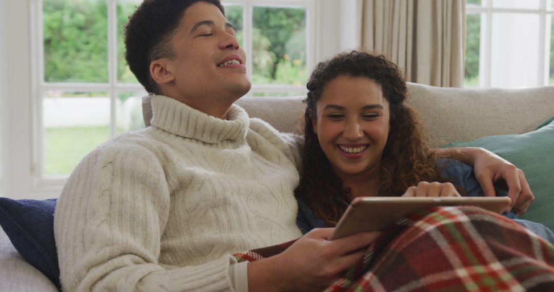Image of happy biracial couple siting on sofa under blanket and using tablet - Free Images, Stock Photos and Pictures on Pikwizard.com