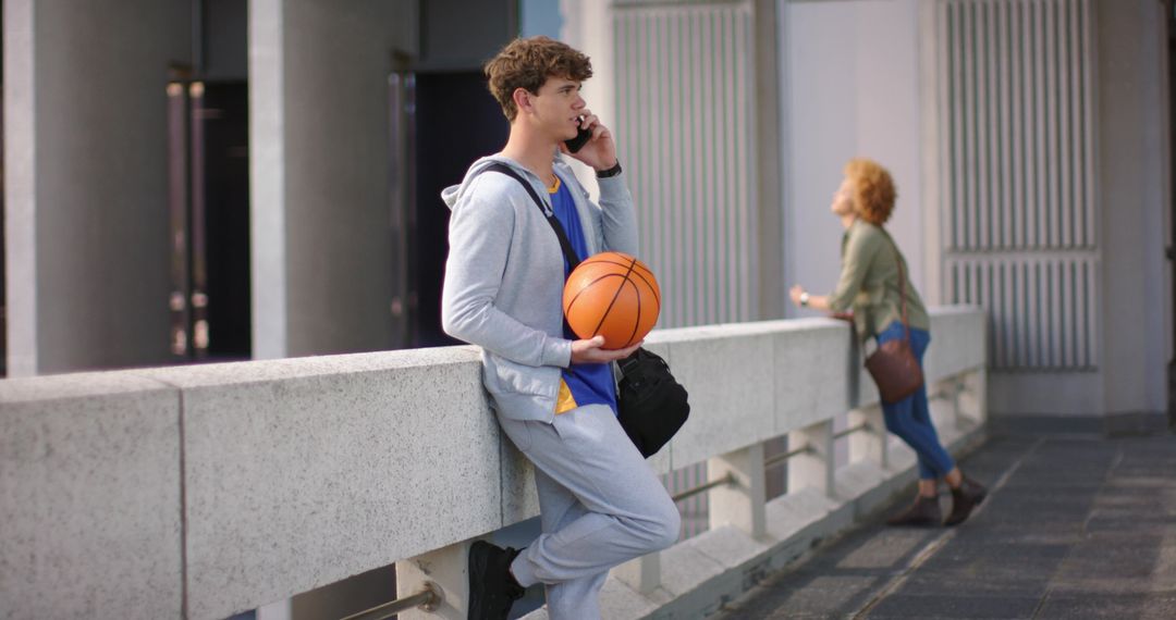 Young man leaning on concrete railing holding basketball while talking on phone in urban walkway