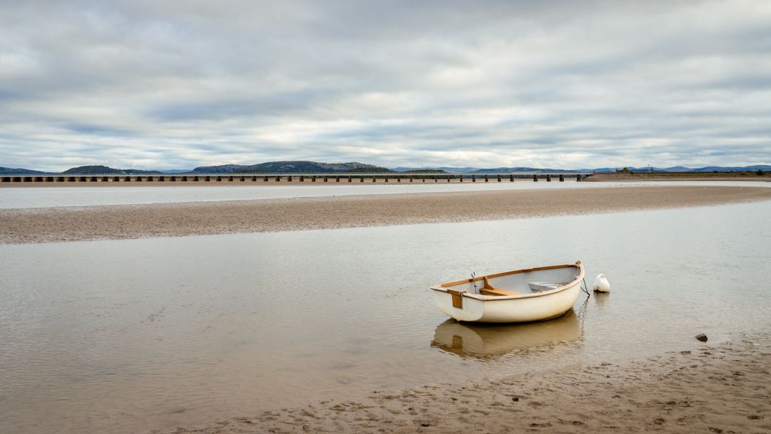 Rowboat resting at low tide on mudflat with buoy and distant bridge under cloudy sky