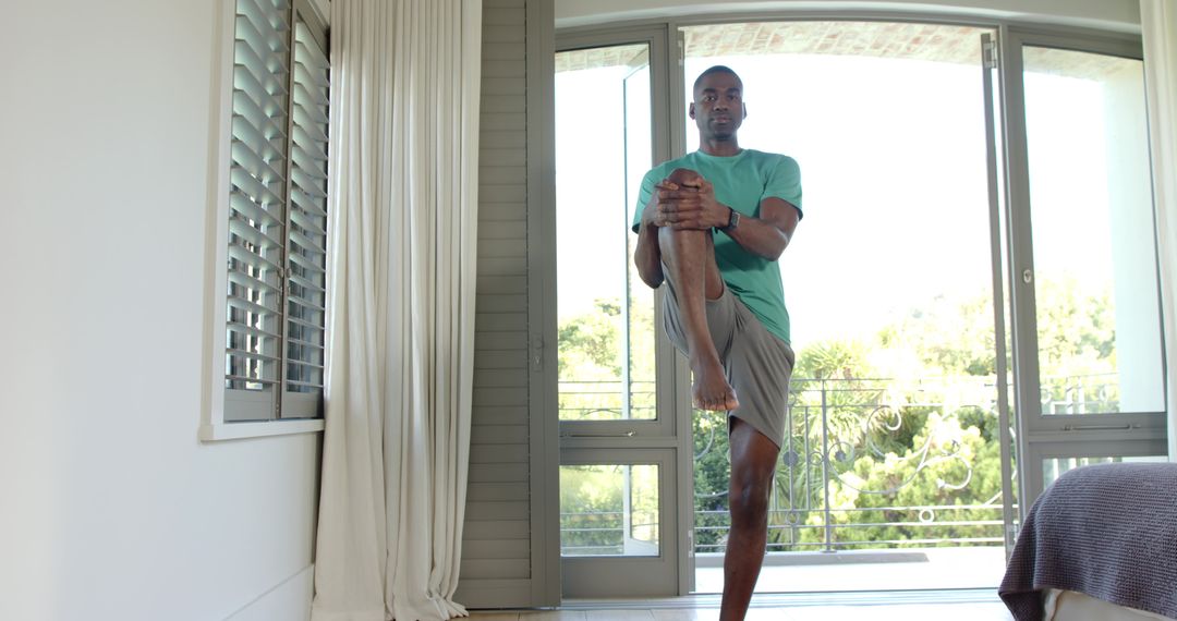 African American man balancing on one leg hugging knee by sliding glass doors at home balcony