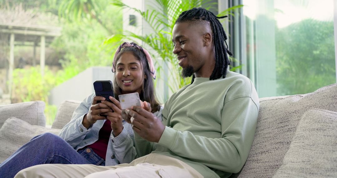Indian Woman and African American Man Making Mobile Payment on Sofa while Smiling