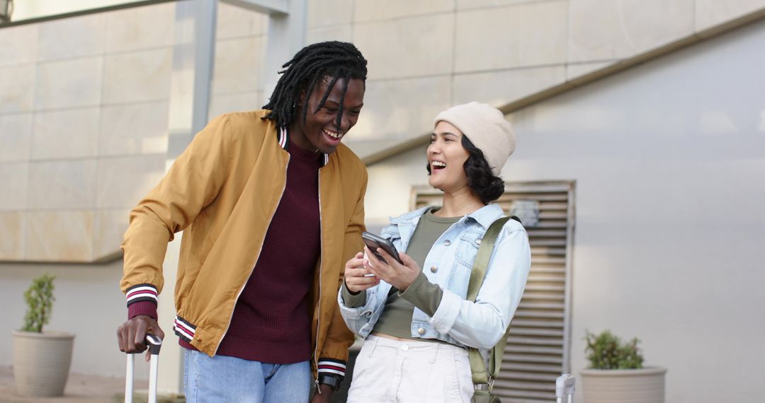 Young diverse couple walking, laughing and sharing smartphone while traveling with luggage