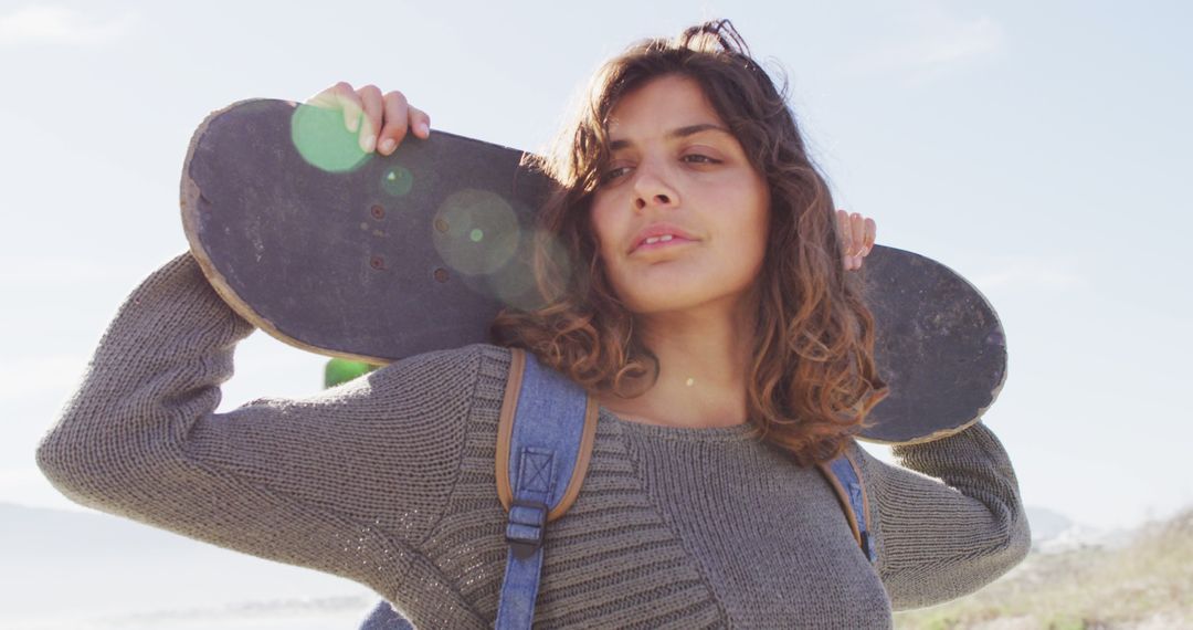 Biracial woman holding skateboard behind head standing in the sun enjoying the view by the sea - Free Images, Stock Photos and Pictures on Pikwizard.com