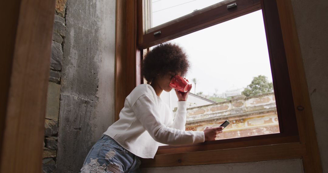 Young Woman Using Mobile Phone and Drinking Coffee by Window - Free Images, Stock Photos and Pictures on Pikwizard.com