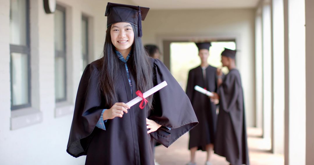 Smiling Chinese graduate holding red-ribboned diploma in school corridor wearing cap and gown