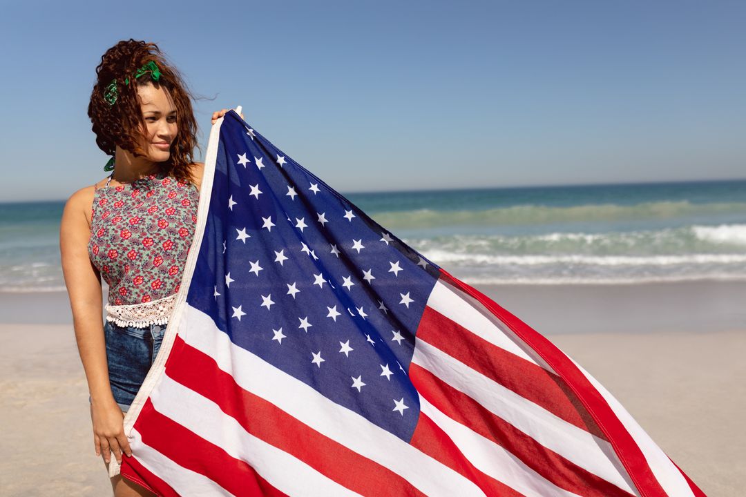 Biracial woman holding American flag at beach, looking thoughtful - Free Images, Stock Photos and Pictures on Pikwizard.com