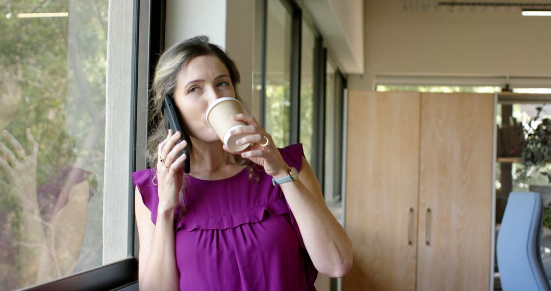 Businesswoman drinking coffee while talking on phone near office window - Free Images, Stock Photos and Pictures on Pikwizard.com