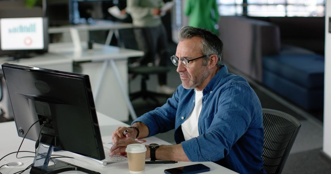Mature professional focusing on computer at open-plan office desk with coffee, smartphone