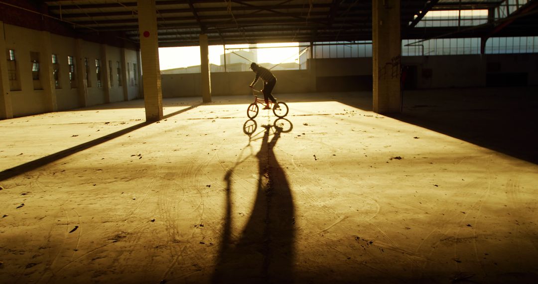 Cyclist Riding in Abandoned Industrial Warehouse at Sunset Casting Long Shadow - Free Images, Stock Photos and Pictures on Pikwizard.com