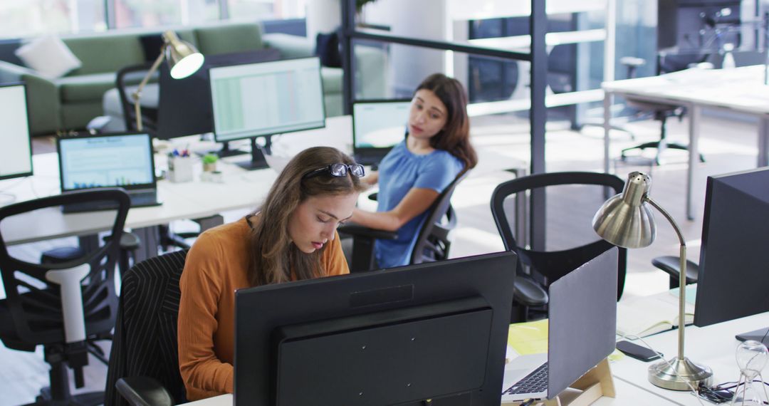 Two caucasian businesswomen sitting at desks using computers turning around to talk to each other - Free Images, Stock Photos and Pictures on Pikwizard.com