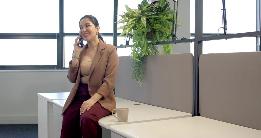 Professional woman talking on smartphone while sitting on desk in modern office with plants