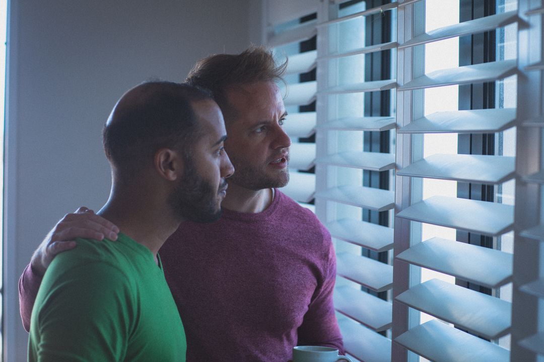 Diverse gay male couple with arms around each other looking out of window through blinds - Free Images, Stock Photos and Pictures on Pikwizard.com
