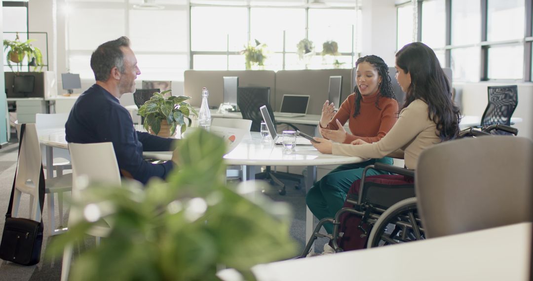 Inclusive team collaborating around table in modern office with wheelchair, laptop, tablet