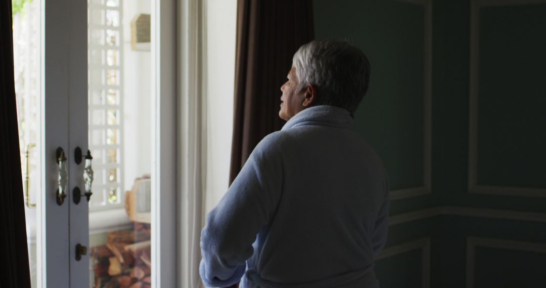 Senior Woman in Bathrobe Looking Out Window in Sunlit Room - Free Images, Stock Photos and Pictures on Pikwizard.com