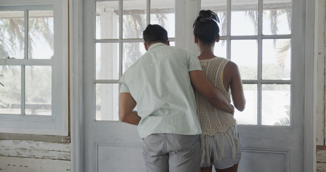 Biracial couple embracing and looking through window in house on beach - Free Images, Stock Photos and Pictures on Pikwizard.com