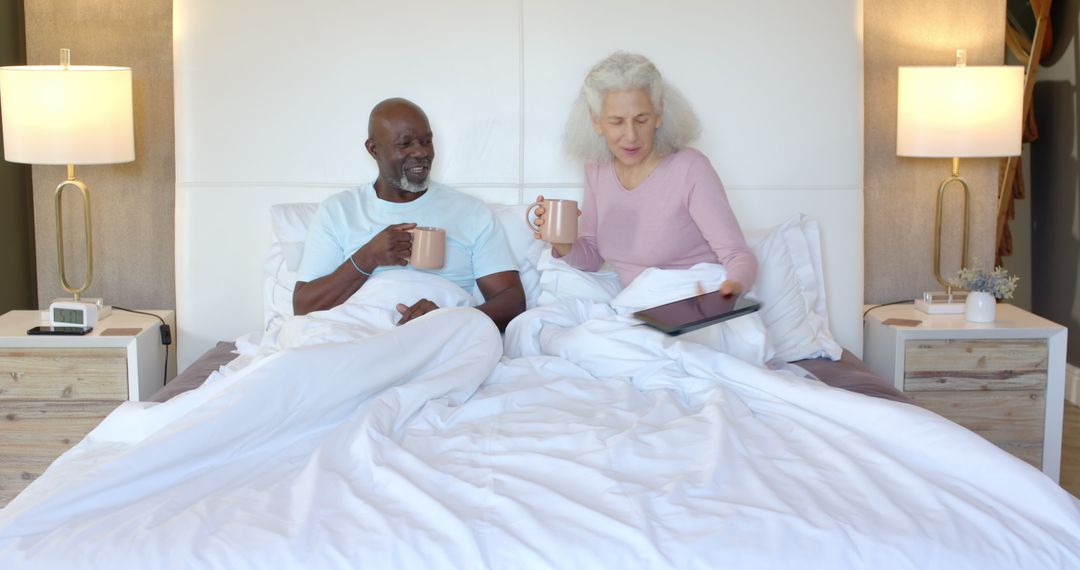 Senior Couple Relaxing in Bed with Mugs and Tablet