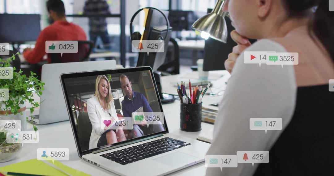 Businesswoman watching virtual meeting on laptop in open office with social notifications