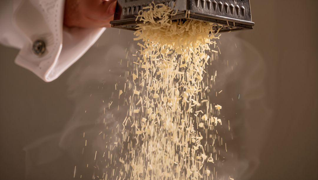 Chef grating hard cheese with box grater, falling shreds forming warm steam and motion