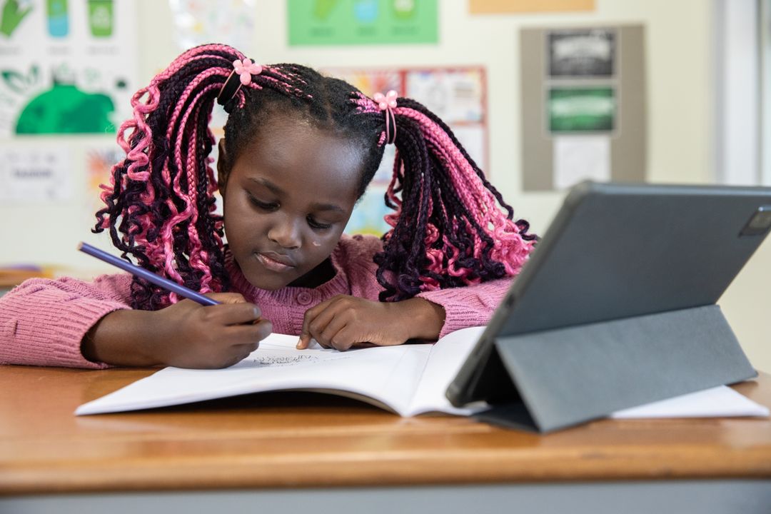 Young Student Writing in Notebook with Tablet at School Desk