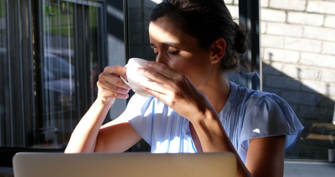 Woman Drinking Coffee while Working on Laptop in Sunlit Café - Free Images, Stock Photos and Pictures on Pikwizard.com