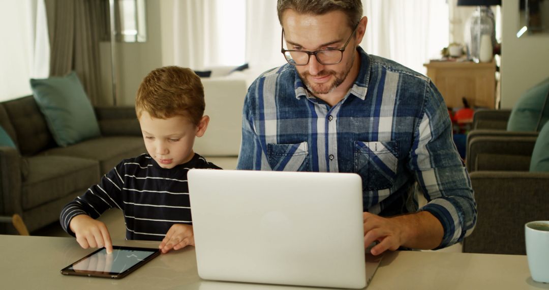 Father Working on Laptop and Son Using Digital Tablet in Living Room - Free Images, Stock Photos and Pictures on Pikwizard.com