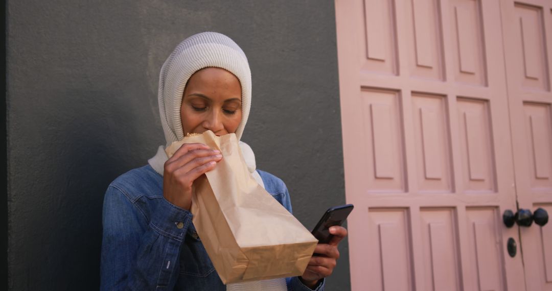 Woman in Hijab Eating from Paper Bag while Using Smartphone Outside - Free Images, Stock Photos and Pictures on Pikwizard.com