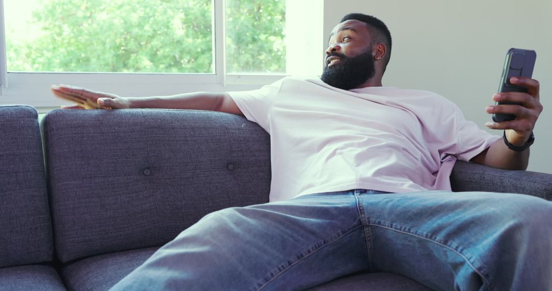 Relaxed Man Lounging on Sofa with Smartphone and Remote Control