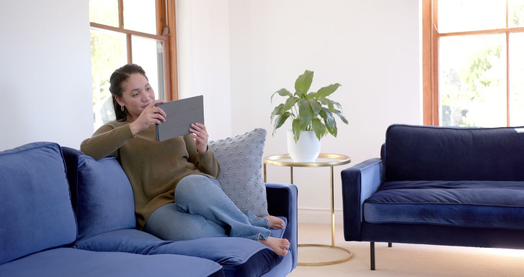 Smiling Woman Relaxing on Blue Sofa Using Tablet in Bright Living Room - Free Images, Stock Photos and Pictures on Pikwizard.com