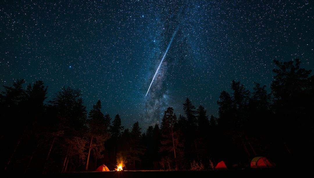 Meteor streaking through Milky Way over campsite with glowing tents and campfire