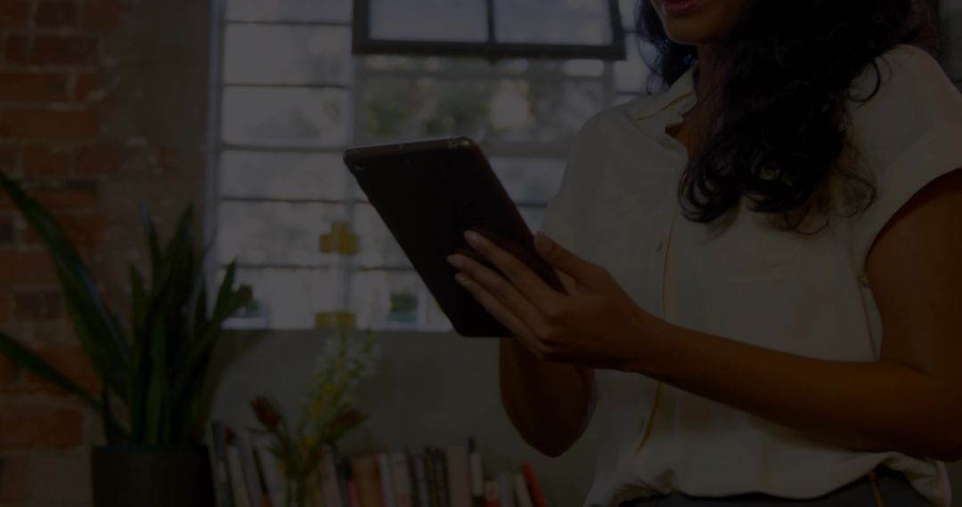 Woman holding tablet tapping screen in cozy home workspace near window and bookshelf