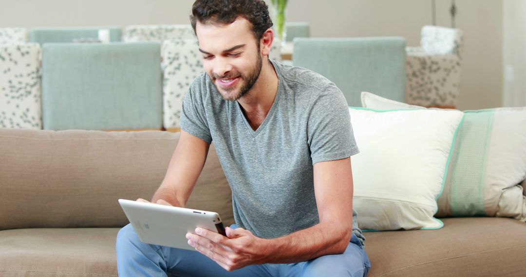 A young Caucasian man is seated on a sofa, engaging with a tablet with a smile, with copy space - Free Images, Stock Photos and Pictures on Pikwizard.com