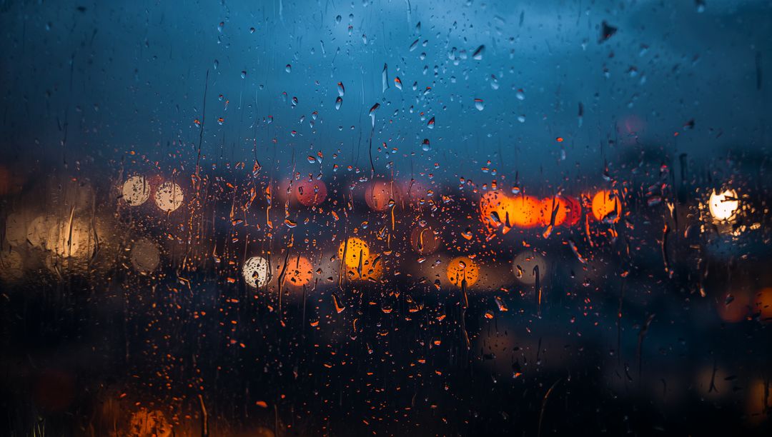 Raindrops streaking on glass window with city lights bokeh at dusk