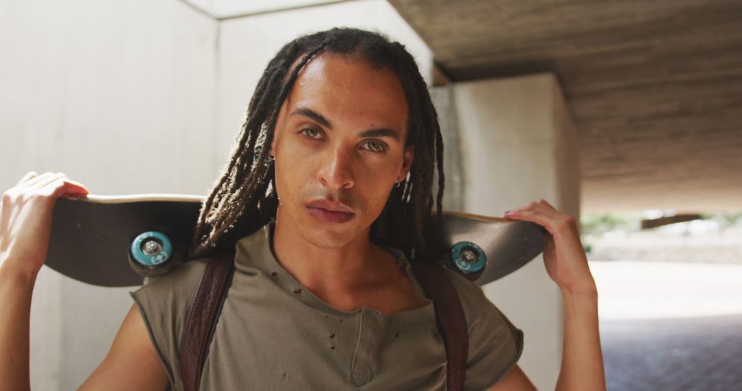 Portrait of fashionable biracial man with dreadlocks holding skateboard and looking at camera - Free Images, Stock Photos and Pictures on Pikwizard.com