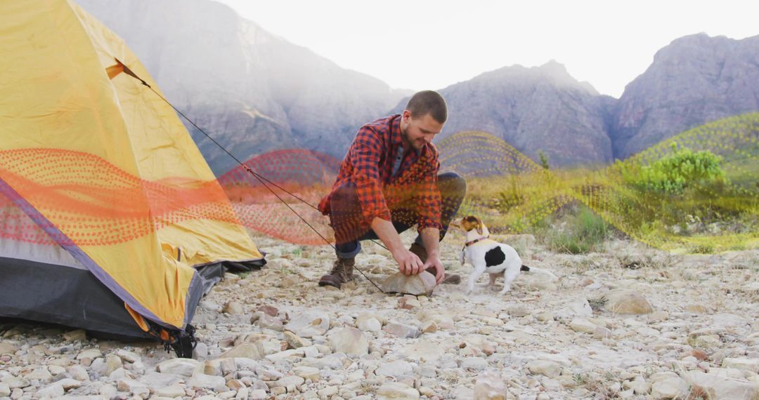 Crouching camper driving tent stake on rocky mountain plain with curious dog observing