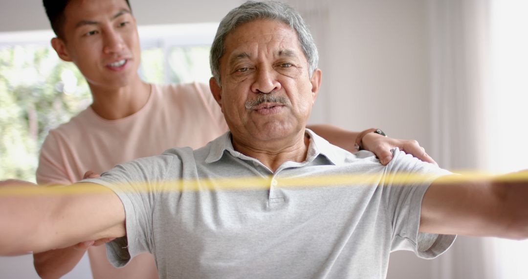 Elderly man exercising with resistant band assisted by caregiver at home - Free Images, Stock Photos and Pictures on Pikwizard.com