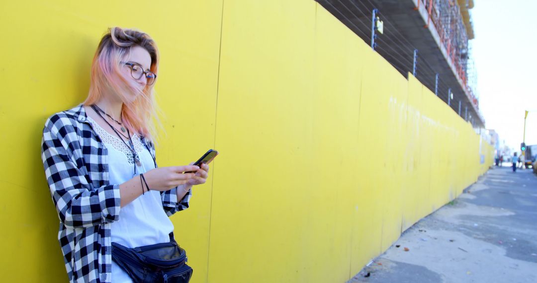 Trendy Young Woman Using Smartphone by Bright Yellow Wall in Urban Area - Free Images, Stock Photos and Pictures on Pikwizard.com