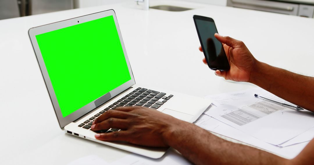 Person Using Laptop with Green Screen and Holding Smartphone at Desk - Free Images, Stock Photos and Pictures on Pikwizard.com