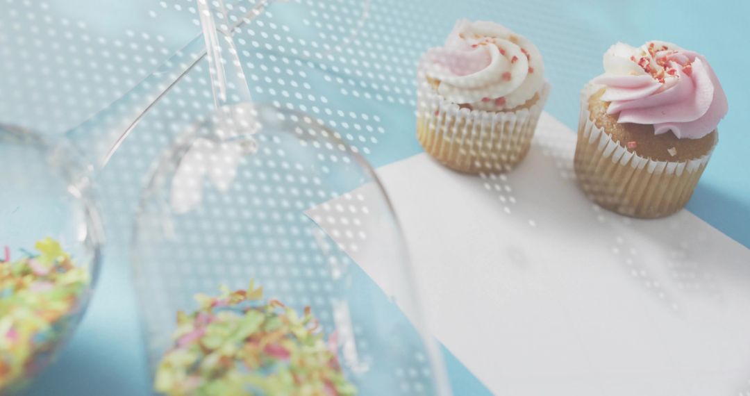 Serving pastel cupcakes with pink frosting and sprinkles on blue tabletop with inverted glasses